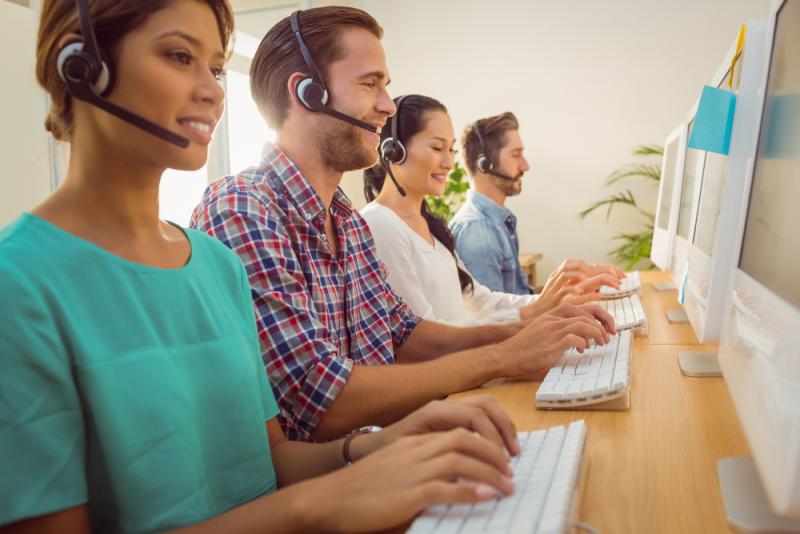 Image of four people wearing headsets, working at computers with keyboards in a modern office, with a plant and sticky notes in the background.