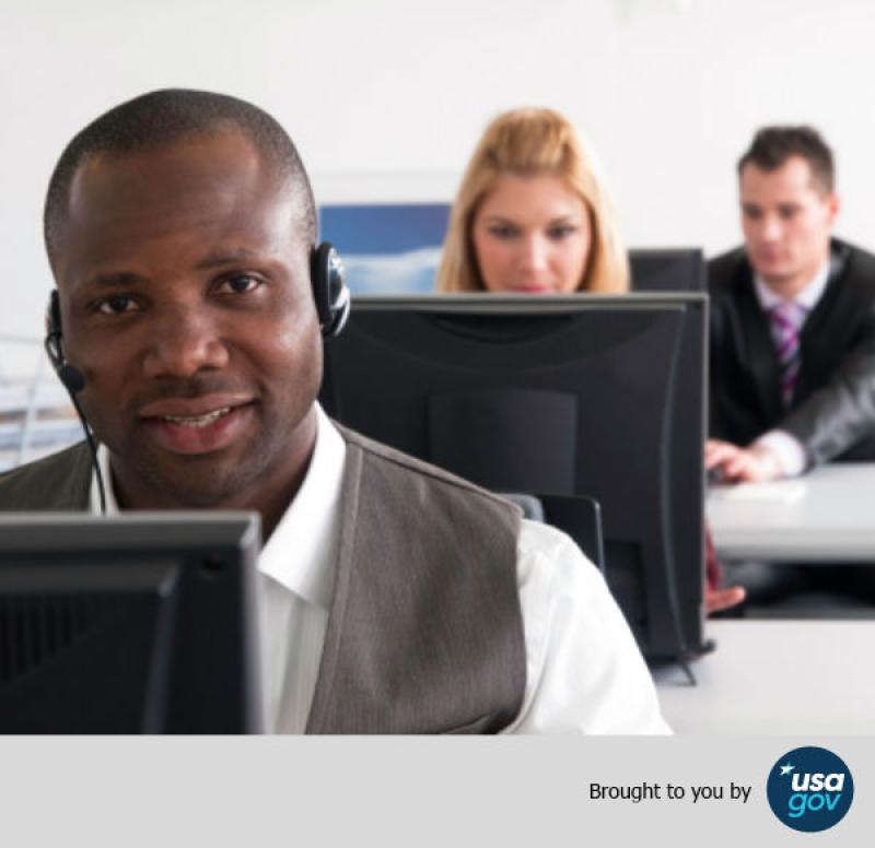 A person wearing a headset at a desk with computer monitors, working in an office with two other individuals in the background, credited to USA.gov.