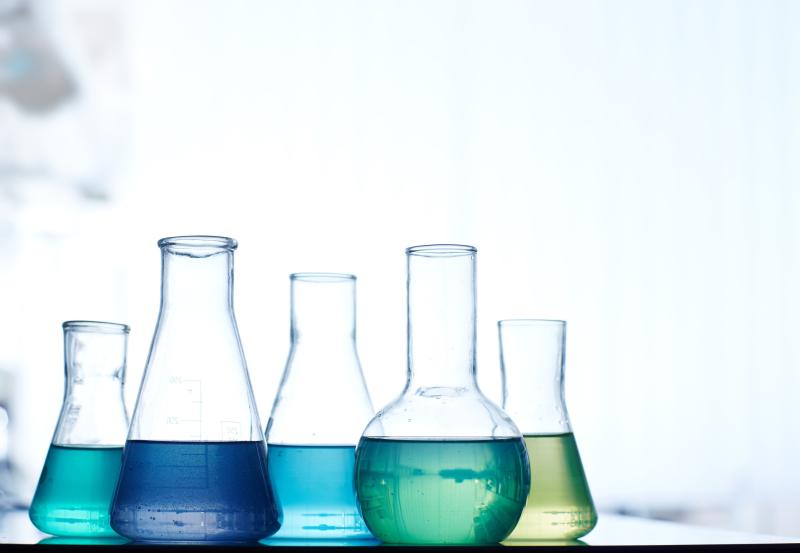A row of laboratory flasks containing colored liquids, including blue, green, and yellow, set against a blurred background.