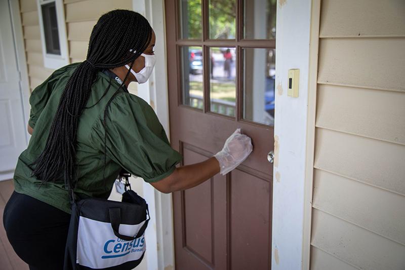 Image of a person in a green shirt and mask knocking on a door, carrying a bag with a "Census" logo, standing on a porch with a beige house exterior.