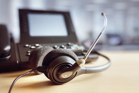 A headset with a microphone resting on a wooden desk, next to a telephone with a small screen, set against a blurred office background.