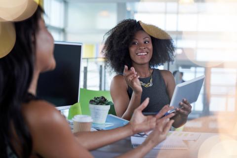 Image of two women in a modern office, one holding a tablet and gesturing, the other at a desk with a computer, coffee cup, and succulent plant.